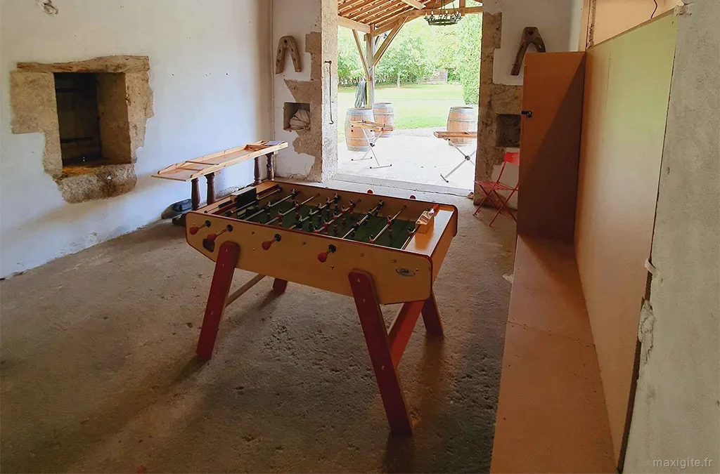 Salle de jeux dans la grange avec baby-foot vintage, murs en pierre, grande ouverture sur la cour et tonneaux en arrière-plan