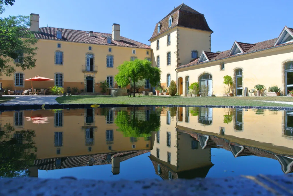 Façade du Château Lacoste en pierre avec tour se reflétant dans le bassin d'eau calme du domaine