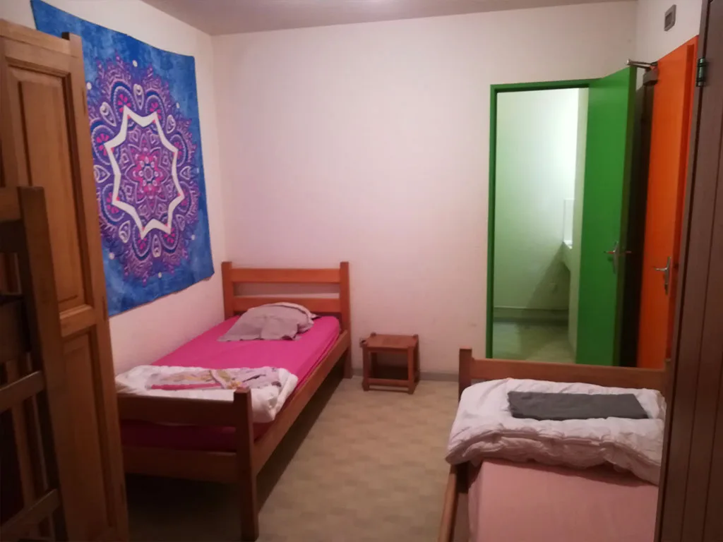 Chambre avec deux lits simples en bois et draps roses, tapisserie bleue décorative au mur, porte verte vers salle d'eau
