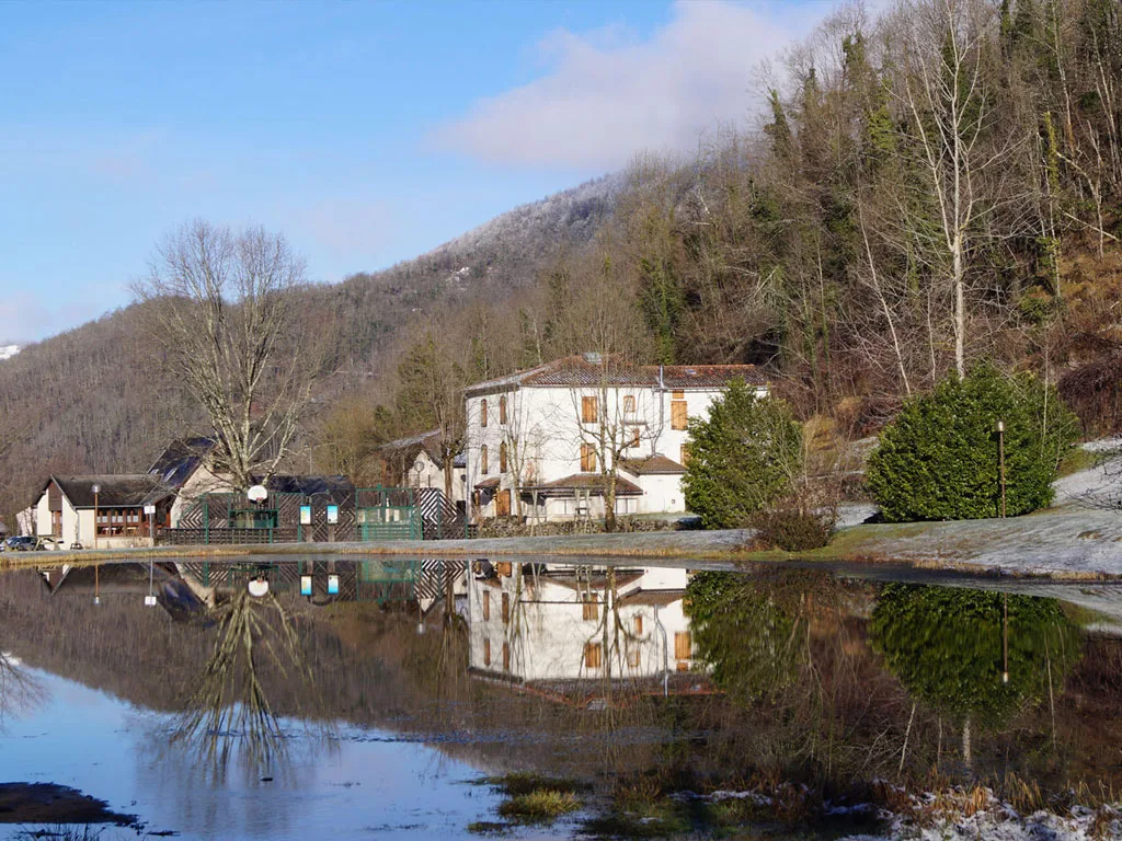 Bâtiments du domaine vus depuis le plan d'eau avec reflets, montagnes pyrénéennes enneigées en arrière-plan sous ciel bleu