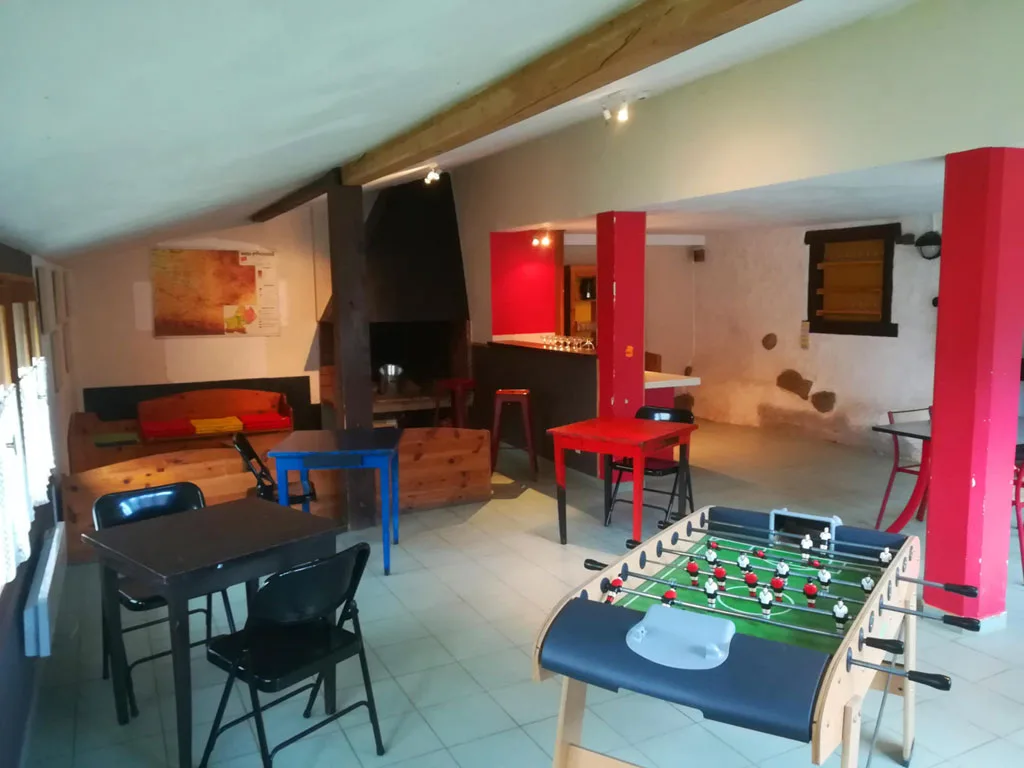 Salle d'activités sous les combles avec poutres apparentes, mur rouge, tables et chaises bleues, éclairage tamisé
