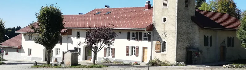Façade blanche du gîte avec toit en tuiles rouges, balcon en bois et jardin verdoyant au pied du Haut-Jura