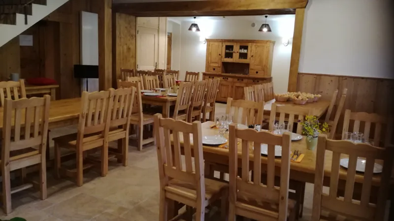 Salle commune avec grande table en bois clair, chaises et vaisselier, prête pour un repas de groupe