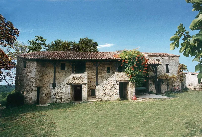Façade en pierre du hameau avec toit de tuiles, cour engazonnée et vigne fleurie grimpant sur la pierre