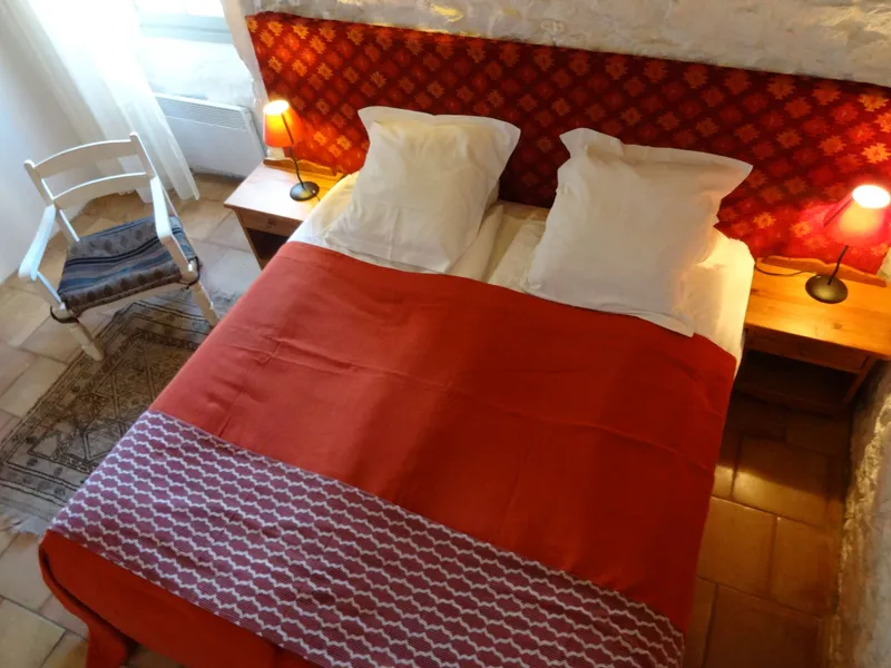 Chambre avec lit double à tête de lit rouge à motifs, chaise blanche et sol en tomettes anciennes