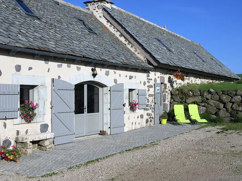 Façade en pierre volcanique du buron avec volets gris, toit de lauzes et terrasse gravillonnée