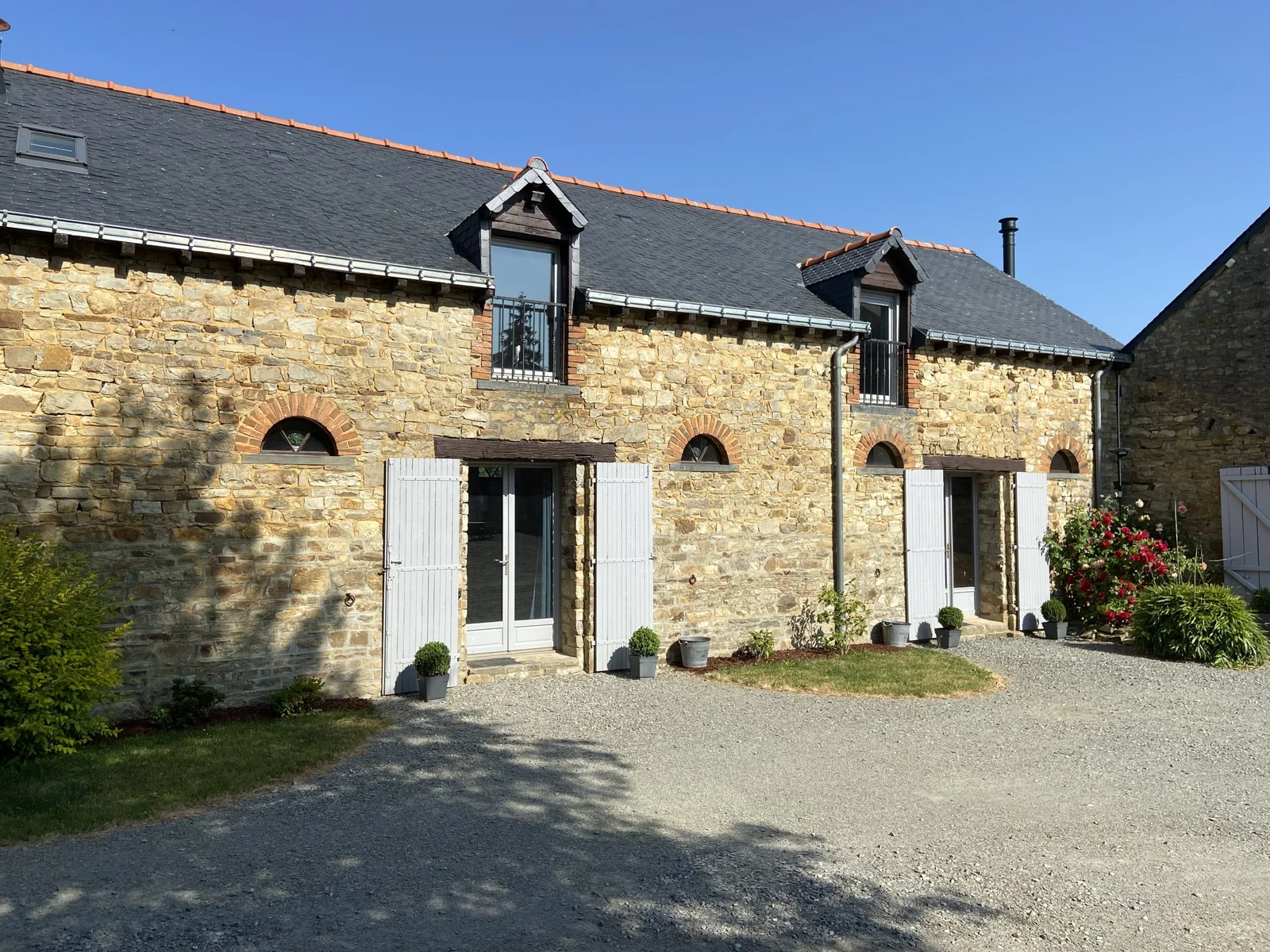 Gîte de groupe Domaine de la Béguinais à Thourie — Façade en pierre du domaine avec toit d'ardoise, lucarnes et allée gravillonnée fleurie, capacité 20 personnes