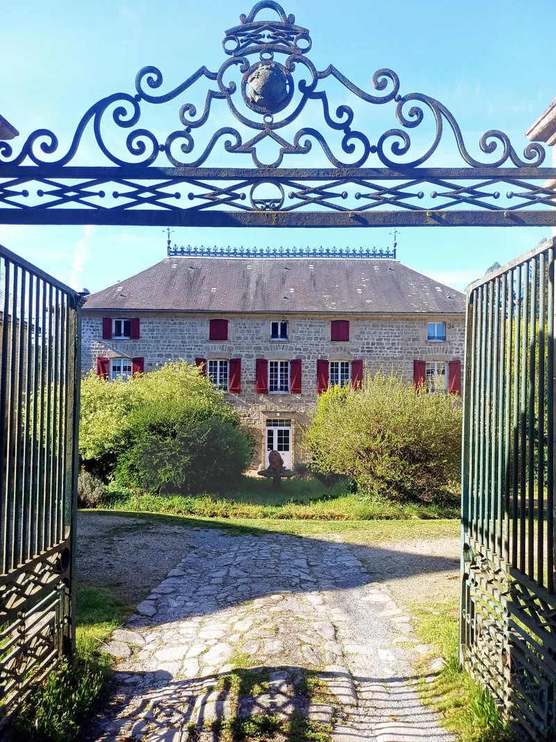 Portail en fer forgé ouvrant sur la façade en pierre blonde du manoir et son parc arboré
