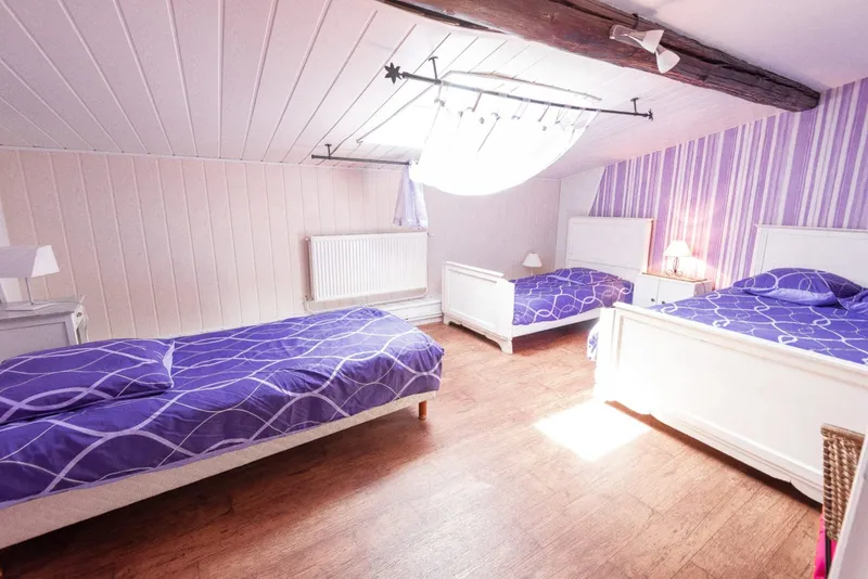 Chambre mansardée avec trois lits simples parés de housses violettes sous velux lumineux