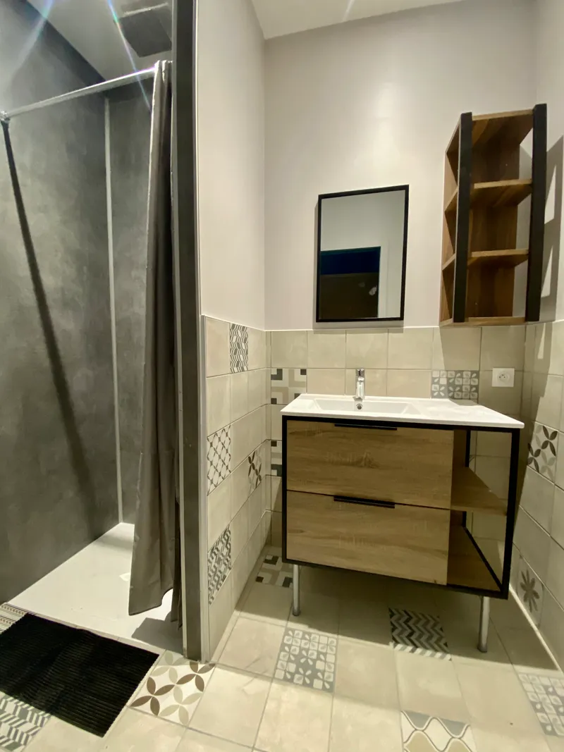 Chambre lambrissée avec lit simple à couverture rouge et couloir carrelé menant aux sanitaires