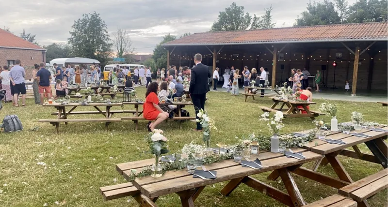 Réception en plein air sous la grange ouverte, tables en bois fleuries et invités attablés