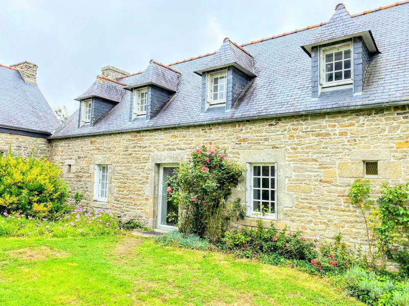 Façade en pierre du gîte avec lucarnes en ardoise, rosiers roses et jardin fleuri