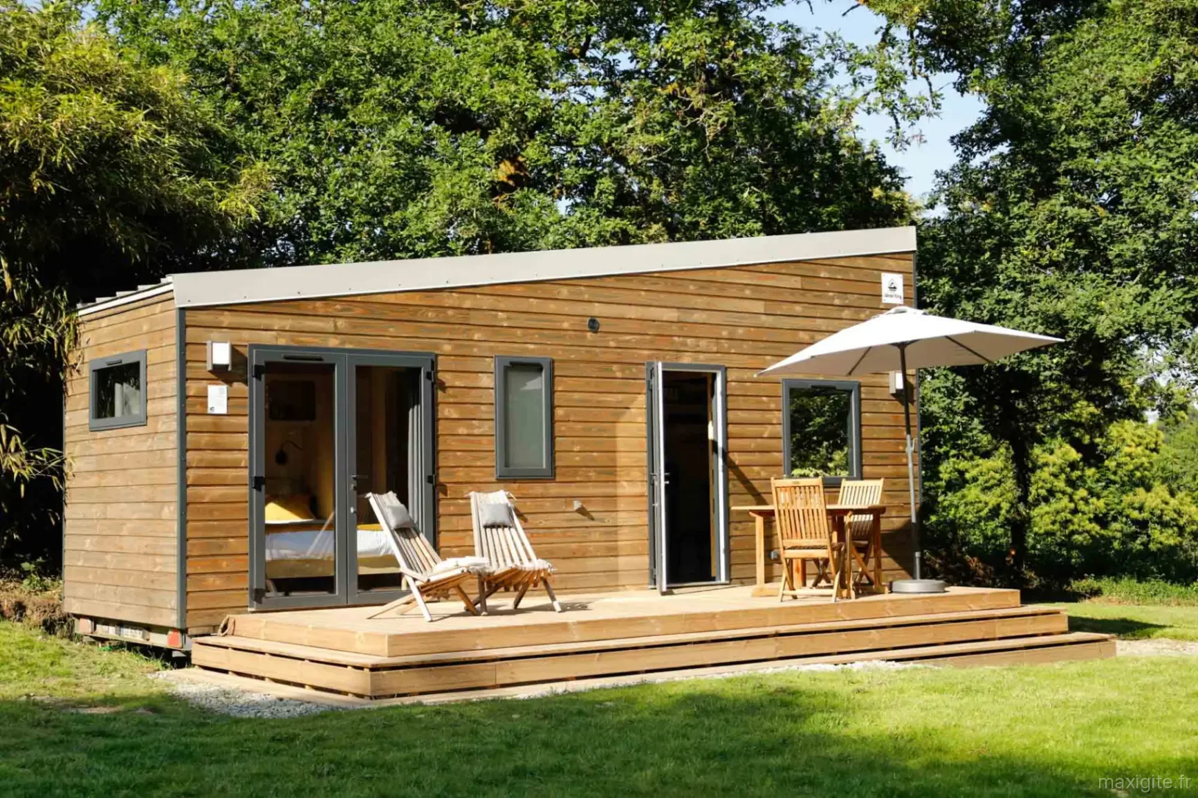 Tiny house Douglas avec large terrasse en bois, chaises longues et parasol face à la forêt