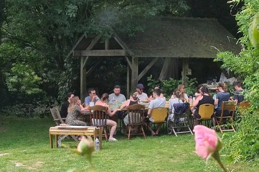 Repas de groupe en plein air sous l'auvent en bois, une vingtaine de convives réunis autour de grandes tables