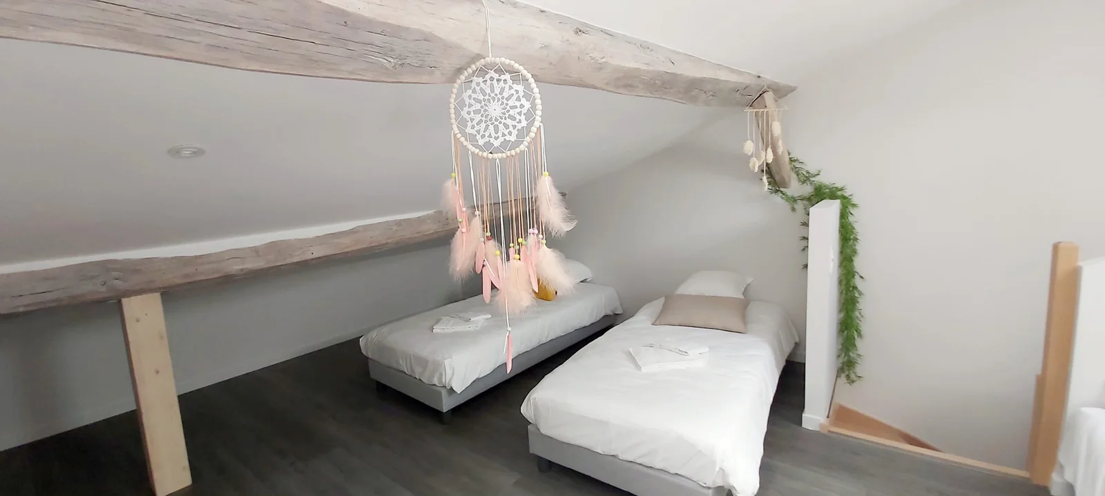 Chambre mansardée avec attrape-rêves à plumes roses suspendu à la poutre et deux lits simples habillés de blanc et gris