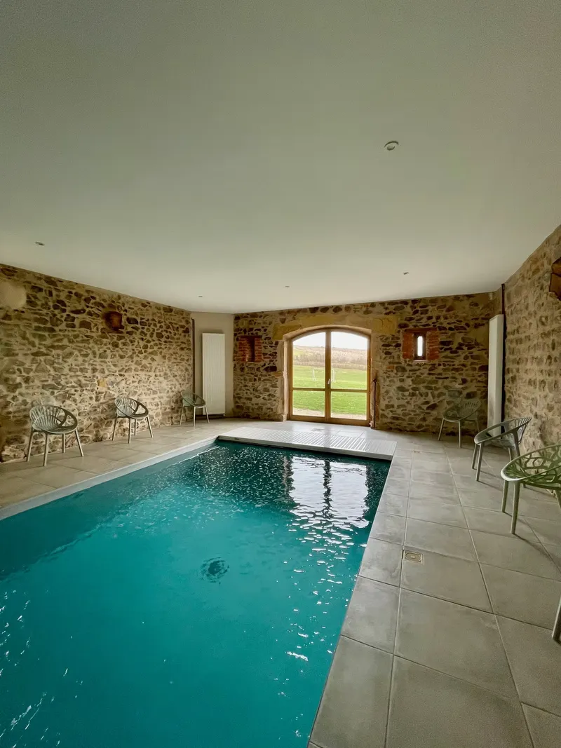Piscine intérieure chauffée entourée de murs en pierre, avec grande baie vitrée donnant sur la campagne