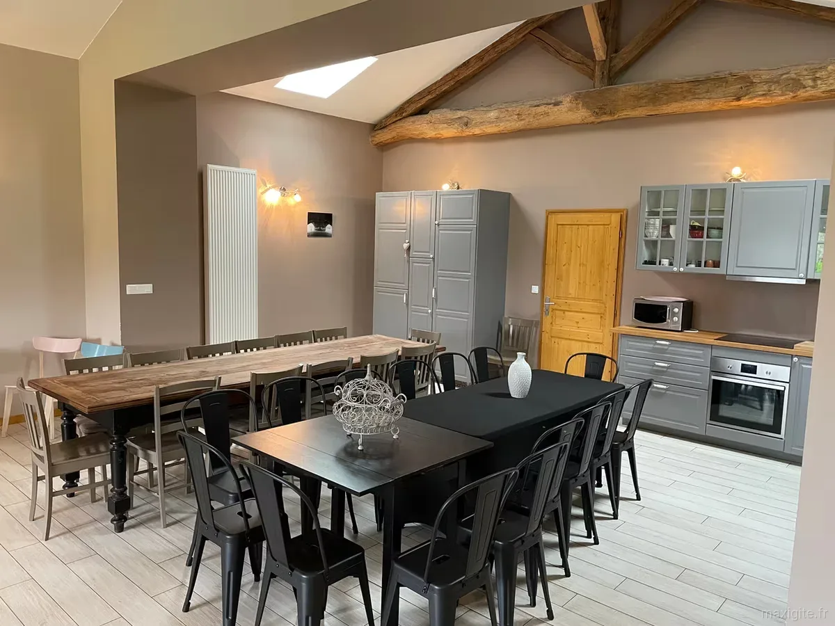 Salle commune avec deux grandes tables, chaises noires et grises, et cuisine ouverte équipée en fond
