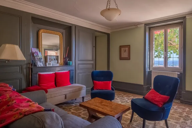 Salon cosy aux murs gris-vert avec fauteuils bleu marine, coussins rouges et miroir doré ancien