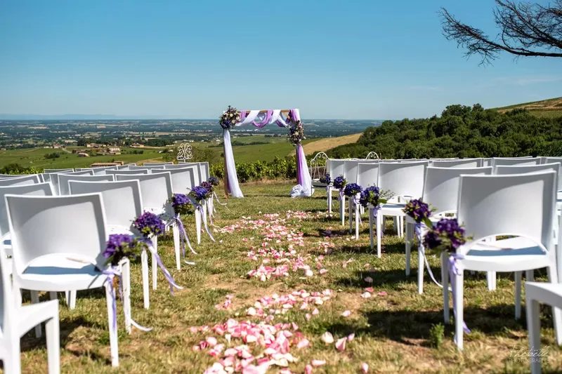 Allée cérémonielle extérieure bordée de chaises blanches et de bouquets mauves, jonchée de pétales de roses avant la cérémonie