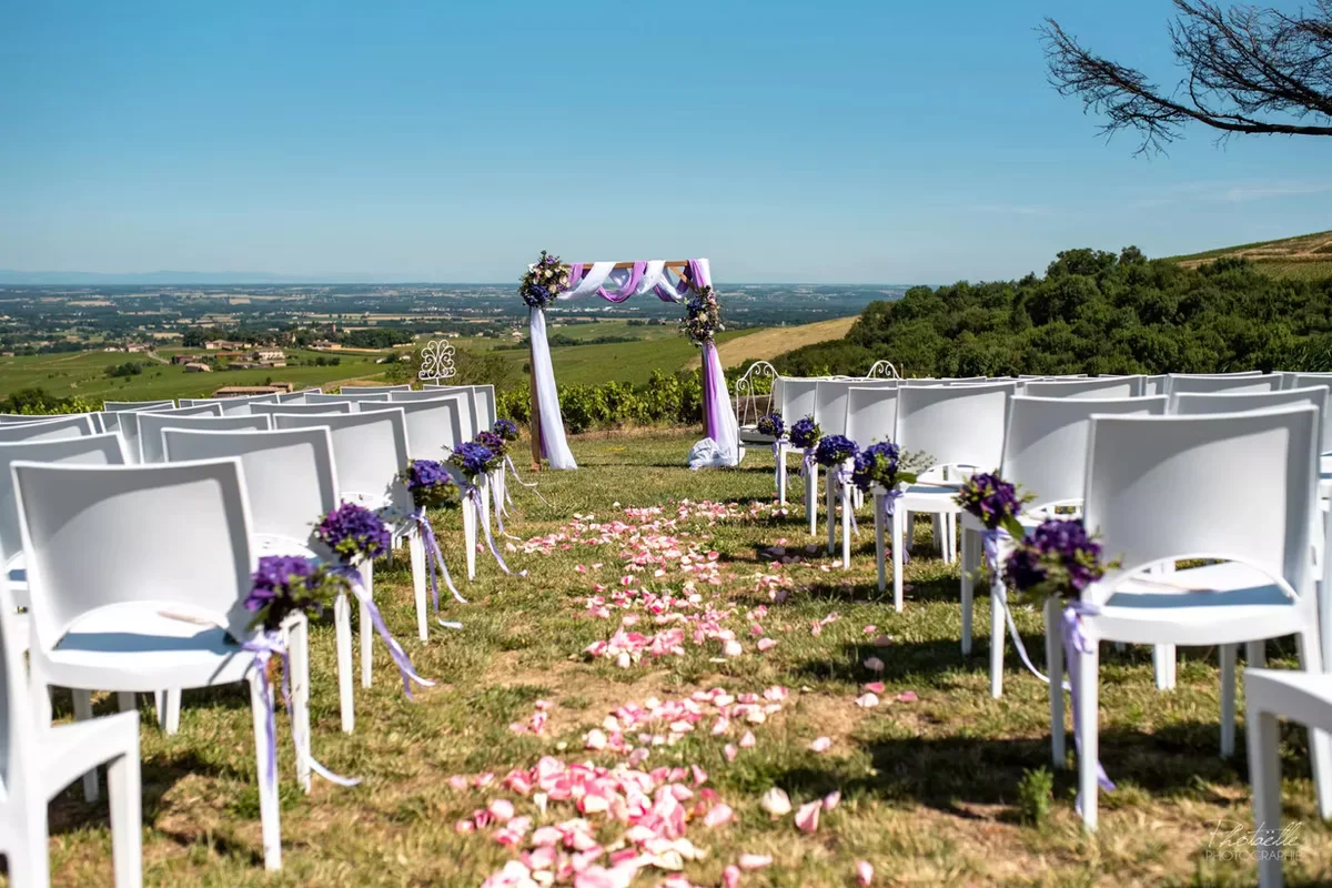 Allée cérémonielle extérieure bordée de chaises blanches et de bouquets mauves, jonchée de pétales de roses avant la cérémonie