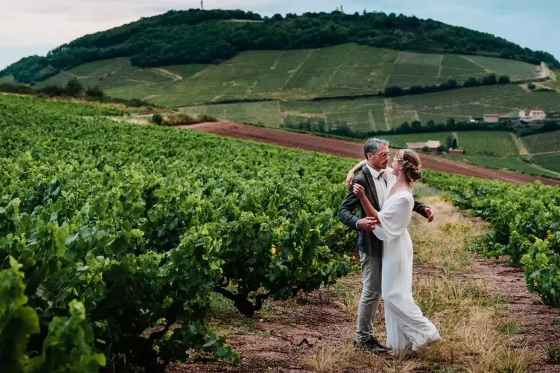 Couple de mariés enlacé au milieu des rangs de vigne, colline du Beaujolais en arrière-plan