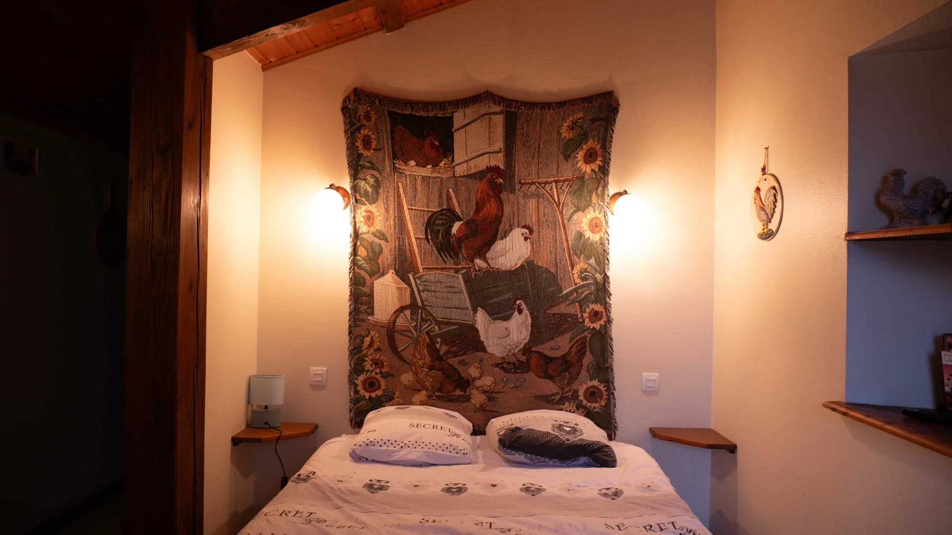 Chambre ornée d'une tapisserie tissée à motif de coq au-dessus du lit, appliques murales allumées et linge fleuri