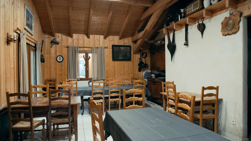 Salle commune rustique aux murs boisés, horloge murale et tables dressées de nappes grises pour les repas