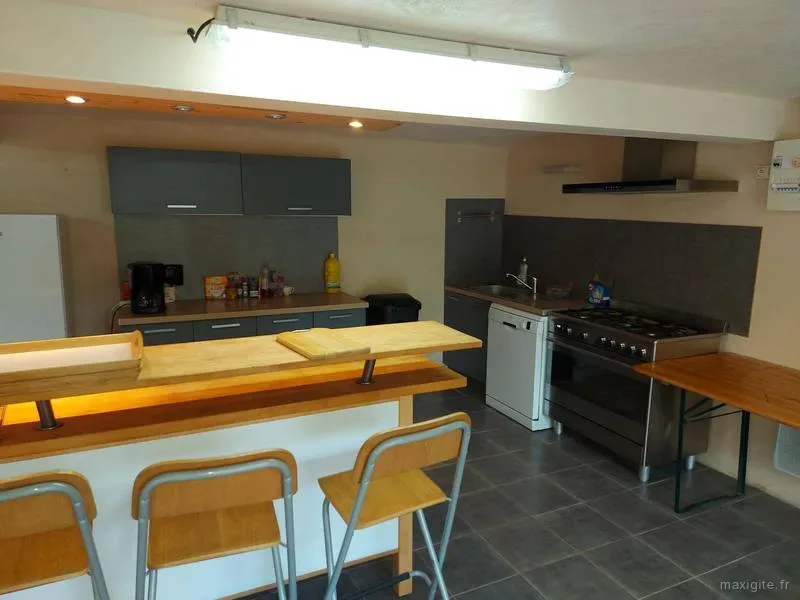 Cuisine commune avec comptoir bar en bois, tabourets, lave-vaisselle et cuisinière inox sous une hotte