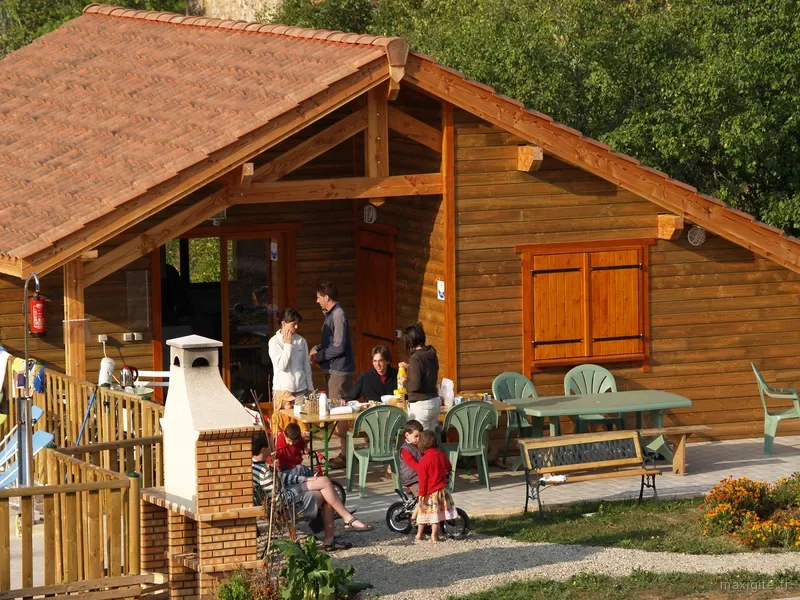 Terrasse d'un chalet en bois avec cheminée barbecue en brique et famille attablée autour d'un repas