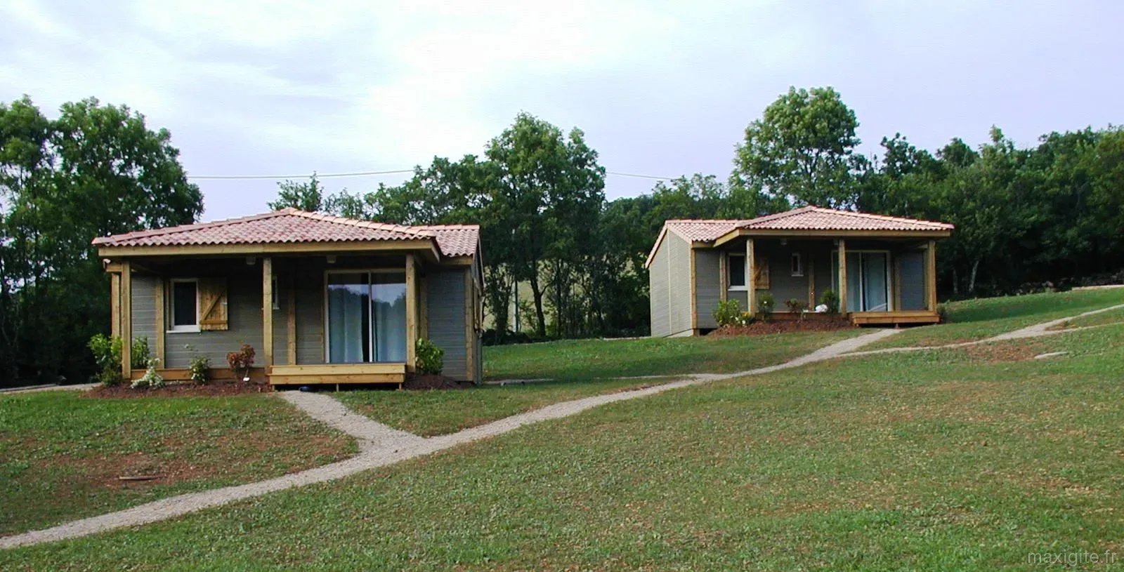 Gîte de groupe Hameau des Celles Basses à Monteils — Deux chalets en bois à toit de tuiles orangées posés sur une pelouse verdoyante, chemin de gravier menant à l'entrée, capacité 30 personnes