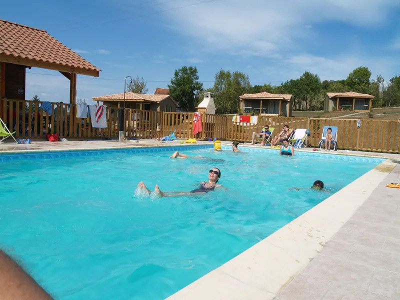 Piscine extérieure animée de baigneurs, entourée d'une clôture en bois et dominée par les toits en tuiles des chalets