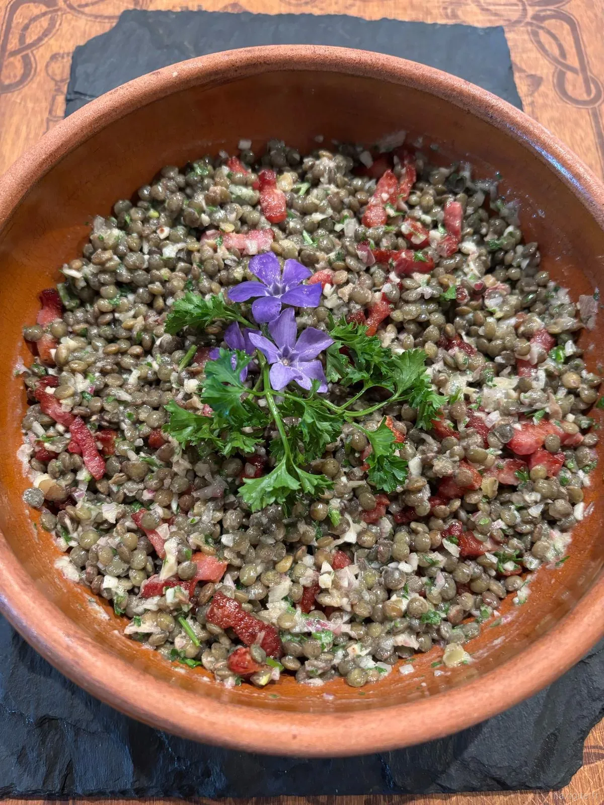 Salade de lentilles maison garnie de tomates et de fleurs comestibles violettes, servie dans un plat en terre cuite