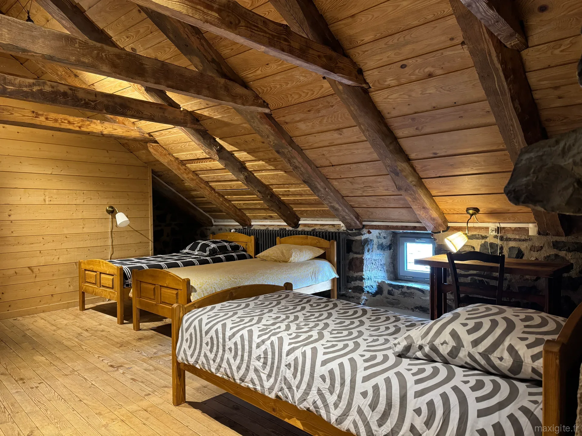 Dortoir sous charpente en bois apparente avec plusieurs lits à motifs géométriques et petit bureau