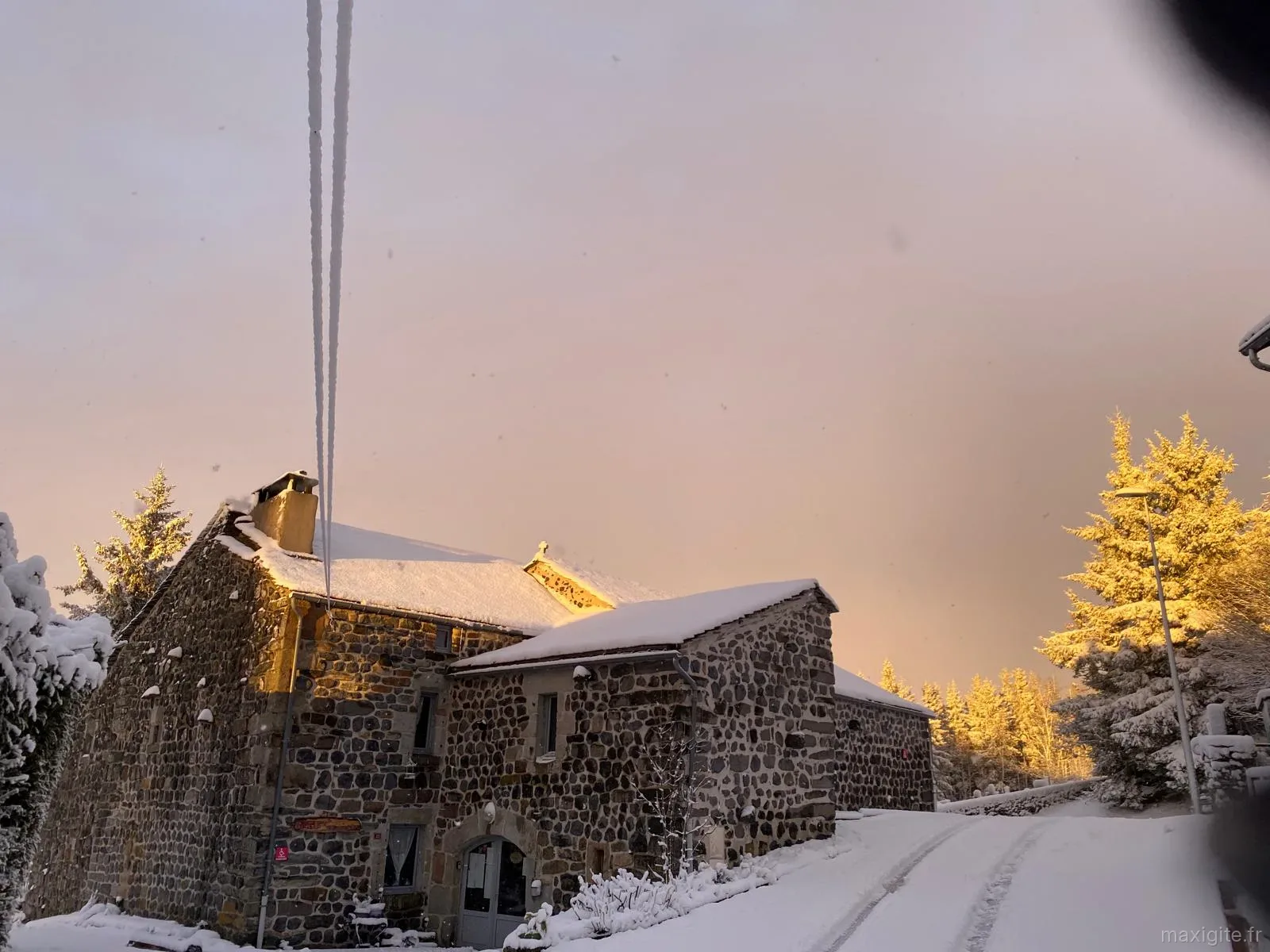 Façade du gîte enneigée au coucher du soleil, toit couvert de neige et sapins dorés par la lumière