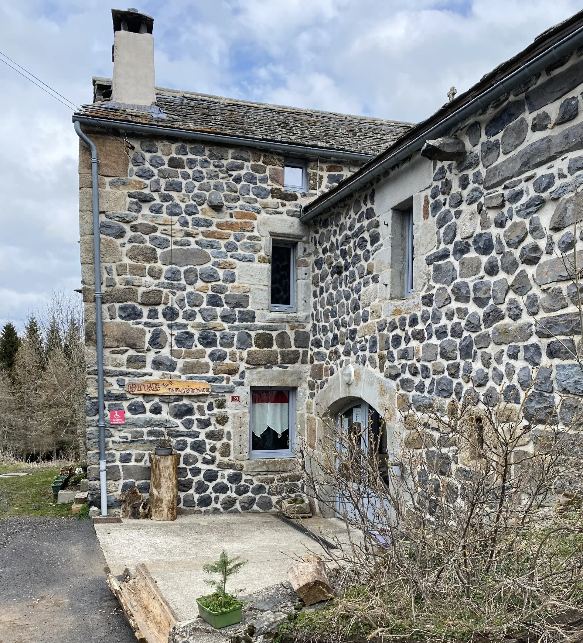 Gîte de groupe Gîte La Traverse à Fay-sur-Lignon — Façade en pierre du gîte avec panneau 'Gîte la Traverse', bûches empilées en décor et porte voûtée bleu ciel, capacité 17 personnes