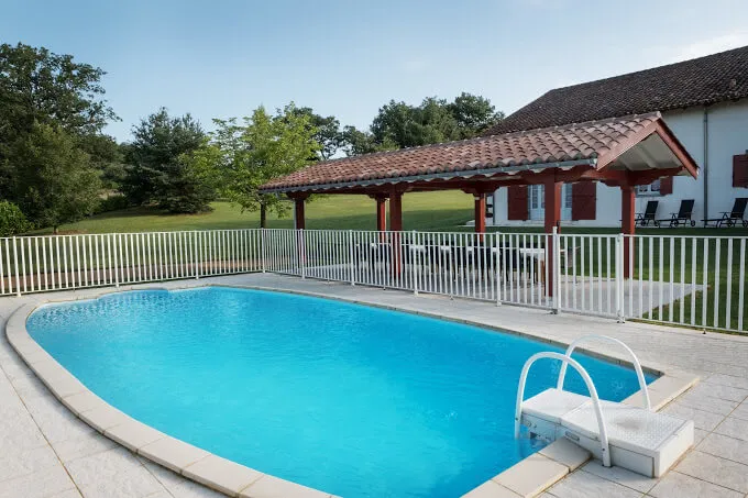 Piscine rectangulaire clôturée de blanc, pergola à toit de tuiles et transats à l'ombre en arrière-plan