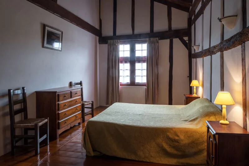 Chambre sous poutres apparentes avec commode en bois ancienne, deux lampes allumées et couvre-lit vert olive