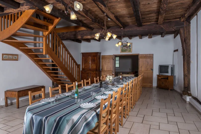 Salle à manger rustique avec escalier en bois tournant, buffet ancien, téléviseur et longue tablée dressée de nappes rayées