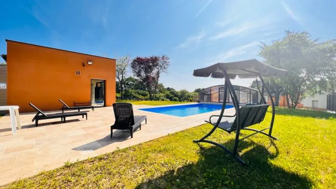 Terrasse extérieure avec transats et balancelle suspendue face à la piscine et à la façade orange du gîte
