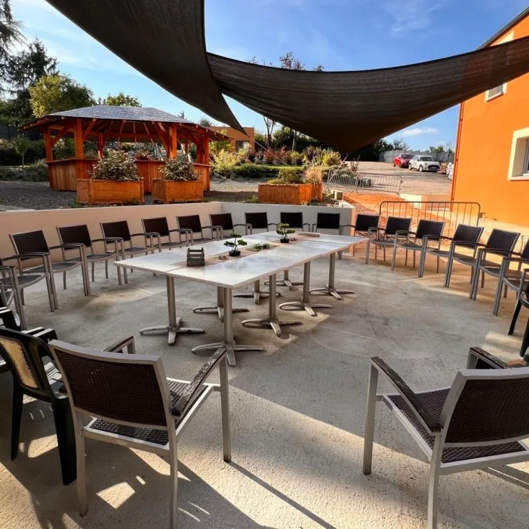 Terrasse couverte de voiles d'ombrage avec chaises disposées en cercle autour de tables, façade orange en fond