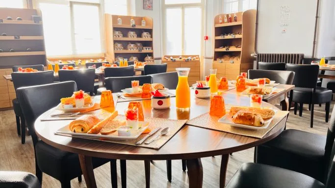 Salle de petit-déjeuner avec table ronde dressée de jus d'orange, viennoiseries et confitures