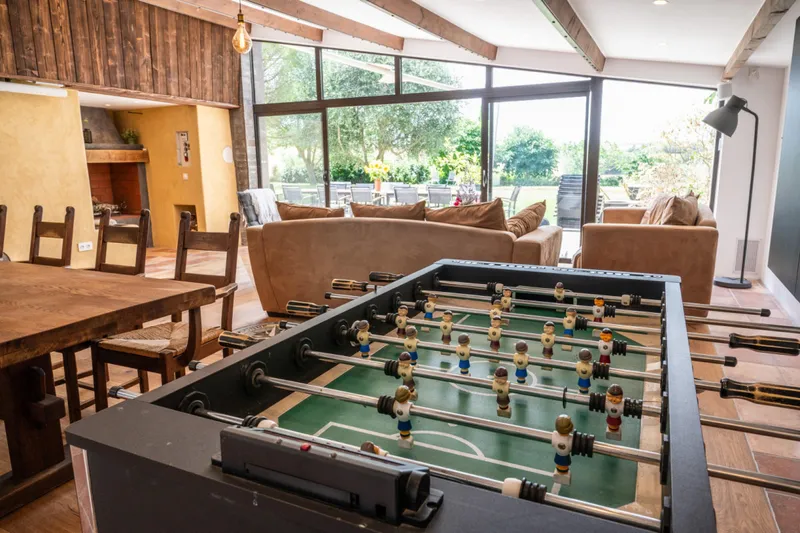 Salon avec baby-foot, grande table en bois, canapes et cheminee, baies vitrees ouvertes sur le jardin