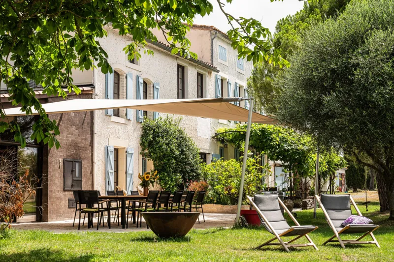Facade en pierre aux volets bleus avec terrasse amenagee sous voile d'ombrage et chaises longues sur la pelouse