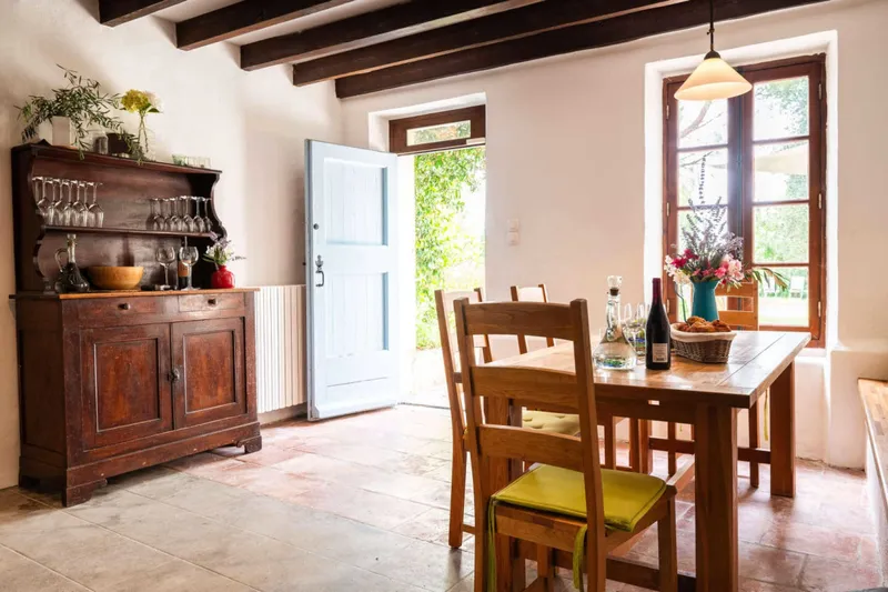 Salle a manger avec vaisselier ancien, poutres apparentes et porte bleue ouverte sur le jardin